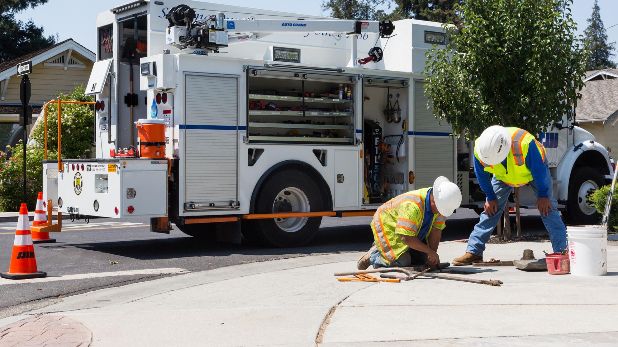 Two service workers examine a hole in the sidewalk with repair truck behind them