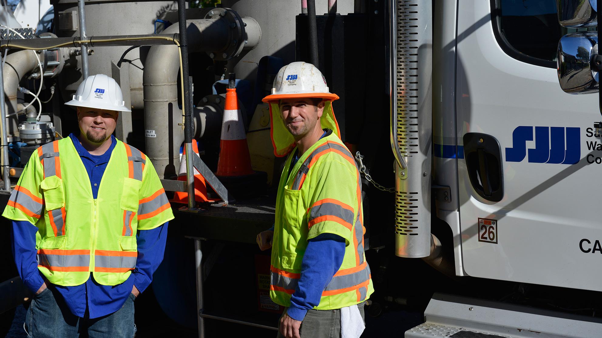 Two workers in hard hats and yellow vests in front of a company utility truck