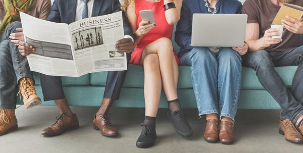 Several people sitting on a couch reading either a newspaper, a book, a smartphone, or a laptop.